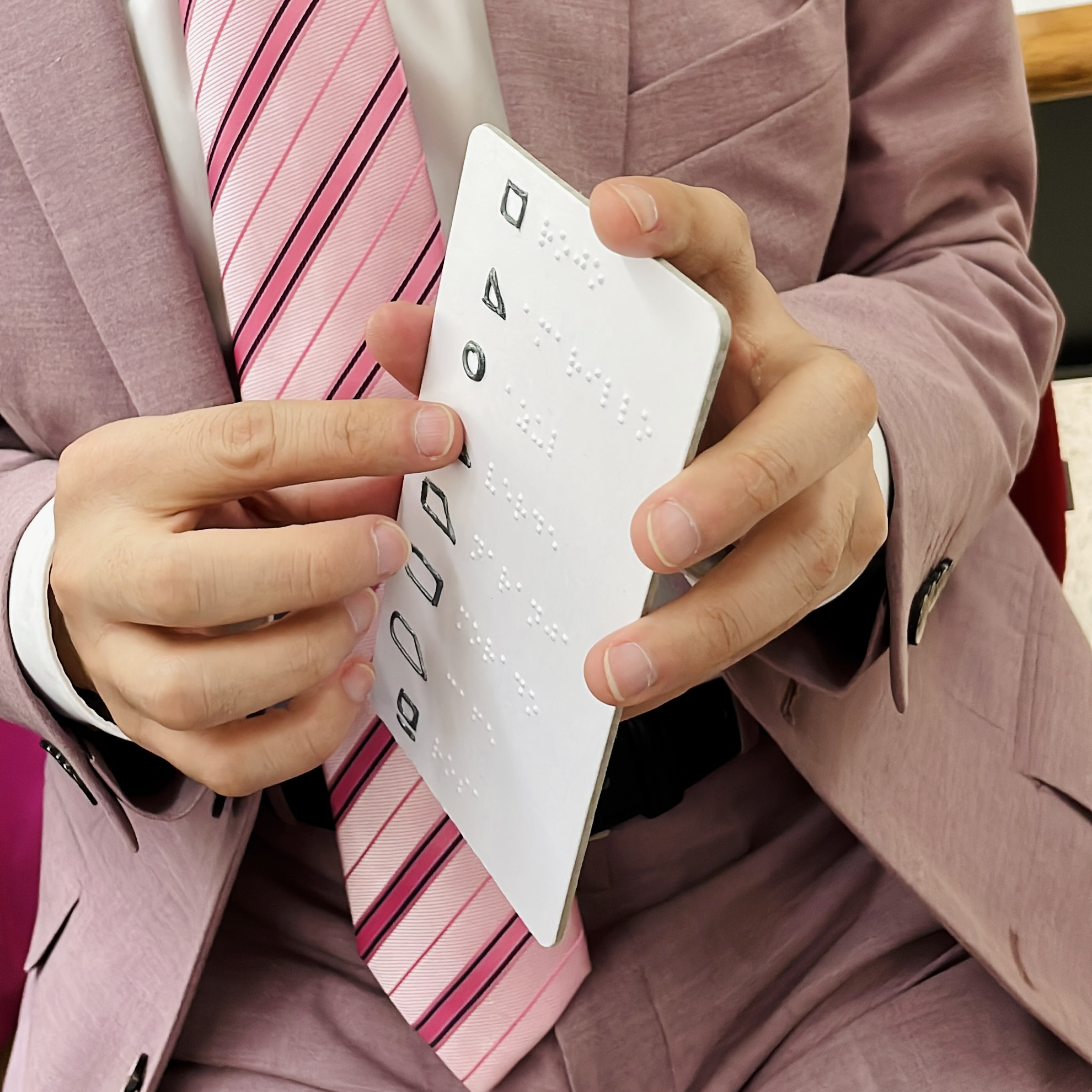 Fotografía a color cuadrada que muestra las manos de una persona tocando una tarjeta vertical con información en braille y una serie de figuras geométricas que representan colores.