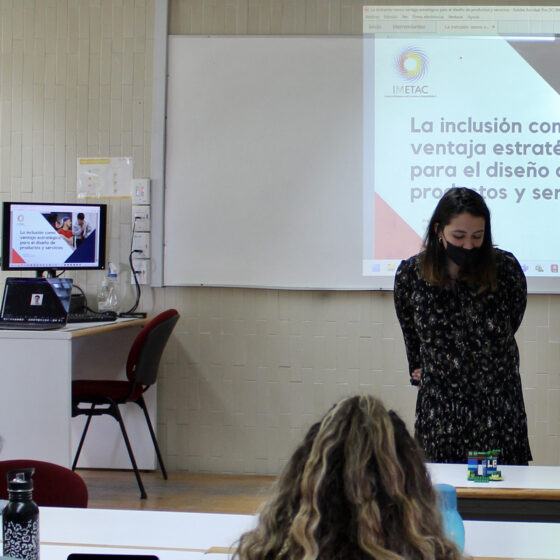 Fotografía a color en posición horizontal, donde se muestra un aula con espectadores de espaldas viendo hacia el frente. En el escenario se puede ver un escritorio y una proyección donde se lee "La inclusión como ventaja estratégica para el diseño de productos y servicios". Una persona está al frente explicando un modelo hecho en una dinámica con la metodología Lego Serious Play.