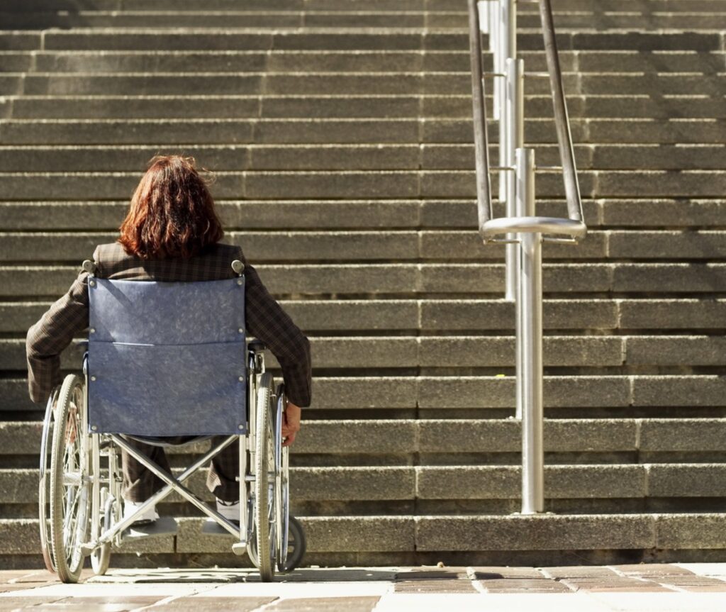 Fotografía horizontal en la que se ve una mujer en silla de ruedas, de espaldas, frente a unas escaleras sin rampa.