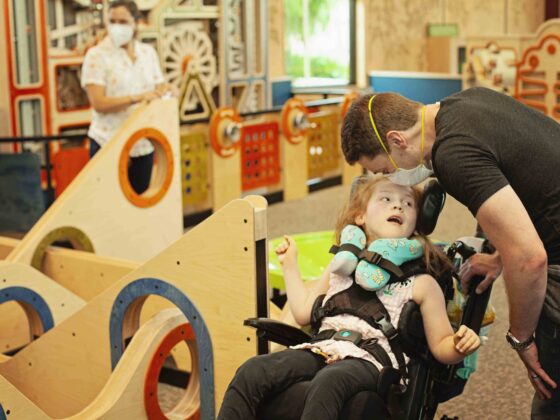 Fotografía de una niña con discapacidad motriz en una silla de ruedas junto a un adulto, recorriendo la sección del museo del zoológico de Palo Alto.