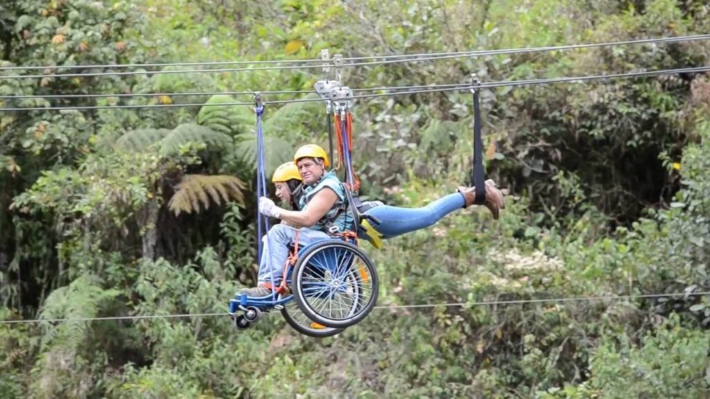 Fotografía horizontal en la que aparecen dos personas deslizándose en una tirolesa. Una de las personas se desliza sobre su silla de ruedas, mientras la otra va con los brazos al frente y piernas colgadas hacia atrás. En el fondo se aprecia el follaje de árboles.