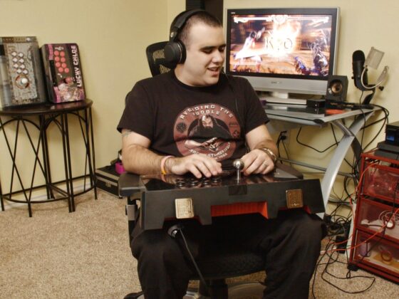 Fotografía de una persona con discapacidad visual jugando videojuegos en una habitación. En su regazo hay una consola con diferentes controles, como botones y palancas. Al fondo hay un escritorio con una computadora encendida y varias consolas de videojuegos.