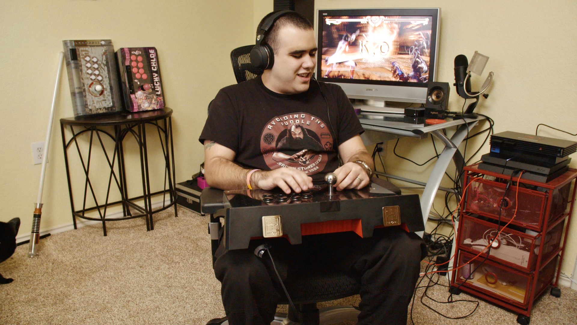 Fotografía de una persona con discapacidad visual jugando videojuegos en una habitación. En su regazo hay una consola con diferentes controles, como botones y palancas. Al fondo hay un escritorio con una computadora encendida y varias consolas de videojuegos.