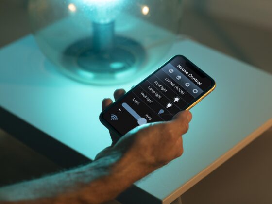 Fotografía de la mano de una persona sosteniendo un dispositivo móvil cerca de una lámpara de mesa. En la pantalla aparecen distintos controles para modificar la intensidad y color de la luz.