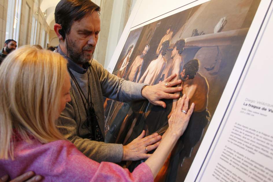 Fotografía de un hombre y una mujer con discapacidad visual sintiendo una obra de arte táctil dentro de un museo. El hombre utiliza audífonos, que están conectados a una audioguía que cuelga de su cuello.