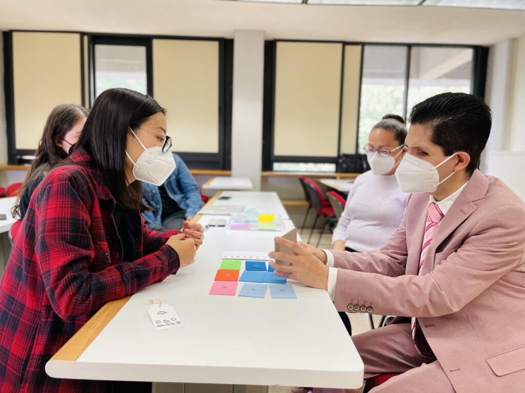 Fotografía horizontal en un salón de clases. Del lado izquierdo al frente aparece la estudiante Harumi Tanaka, quien sobre una mesa muestra una serie de maquetas del sistema táctil para identificar colores. Detrás de ella se ven otros 2 estudiantes. Del lado derecho aparece Cristobal Sanchez palpando los prototipos.