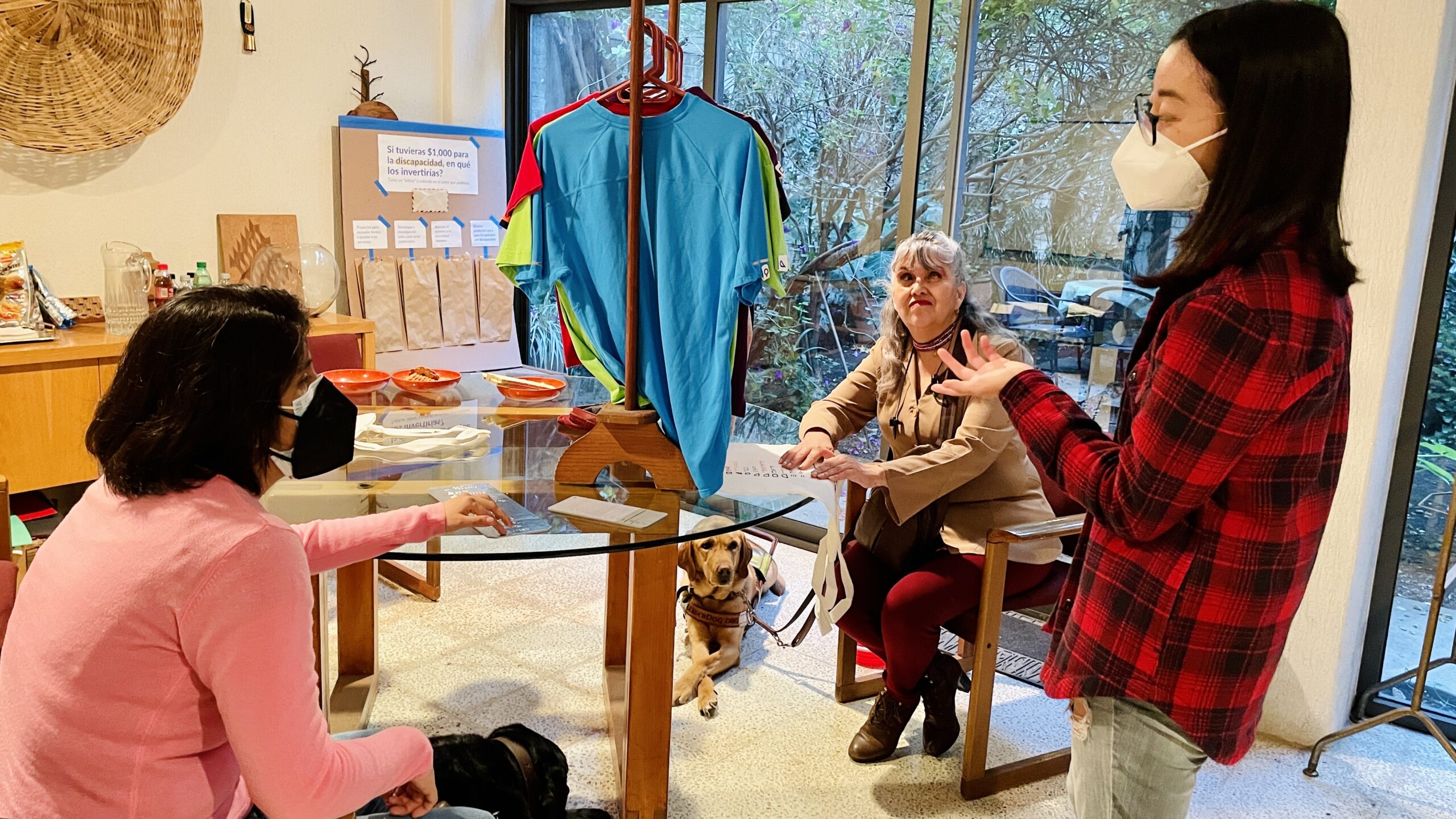 Fotografía horizontal donde dos personas con discapacidad visual están sentadas en una mesa y participan en una simulación de la experiencia de compra de ropa. Se observa una estudiante mostrando un perchero con playeras que incluyen las etiquetas con SIENTOCOLOR cosidas en las mangas. Dos perros guía se asoman por debajo de la mesa.