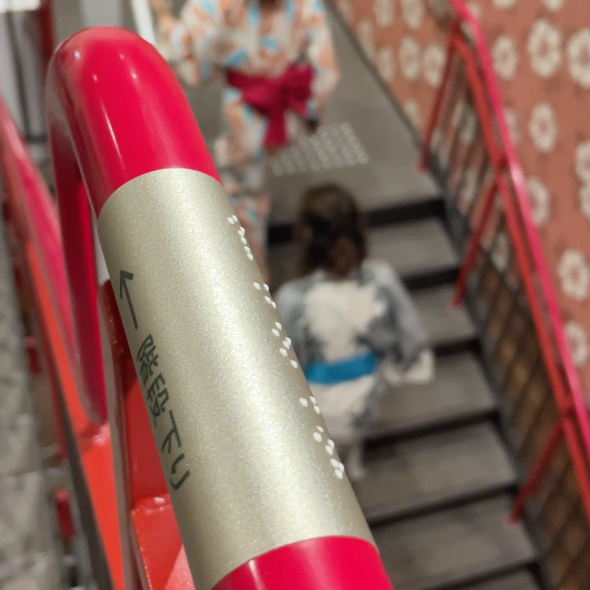 Fotografía del barandal de una escalera que contiene una placa metálica con información en braille. Al fondo se observan dos mujeres con atuendo tradicional japonés subiendo las escaleras, sobre las que hay baldosas podotáctiles.