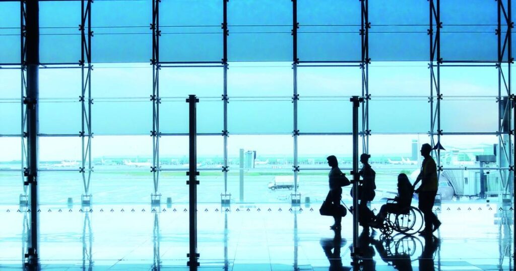 Fotografía horizontal, donde aparece la silueta de cuatro personas caminando en un aeropuerto. Una de ellas es usuaria de silla de ruedas. Al fondo se aprecian ventanales altos con vista hacia la pista de despegue y un avión.