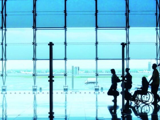 Fotografía horizontal, donde aparece la silueta de cuatro personas caminando en un aeropuerto. Una de ellas es usuaria de silla de ruedas. Al fondo se aprecian ventanales altos con vista hacia la pista de despegue y un avión.