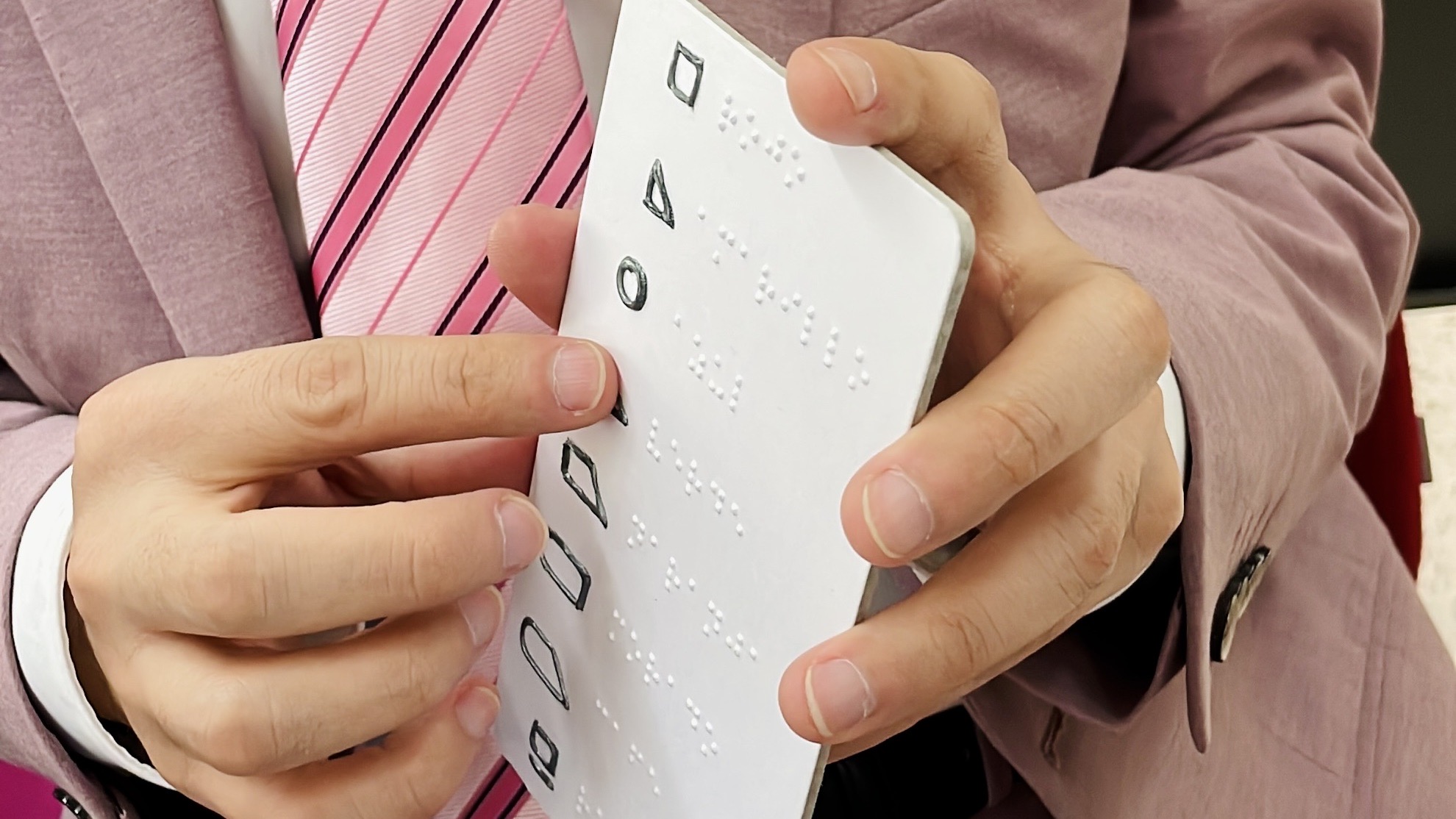Fotografía de las manos de una persona con discapacidad visual tocando una tarjeta vertical con información en braille y una serie de figuras geométricas que representan colores.