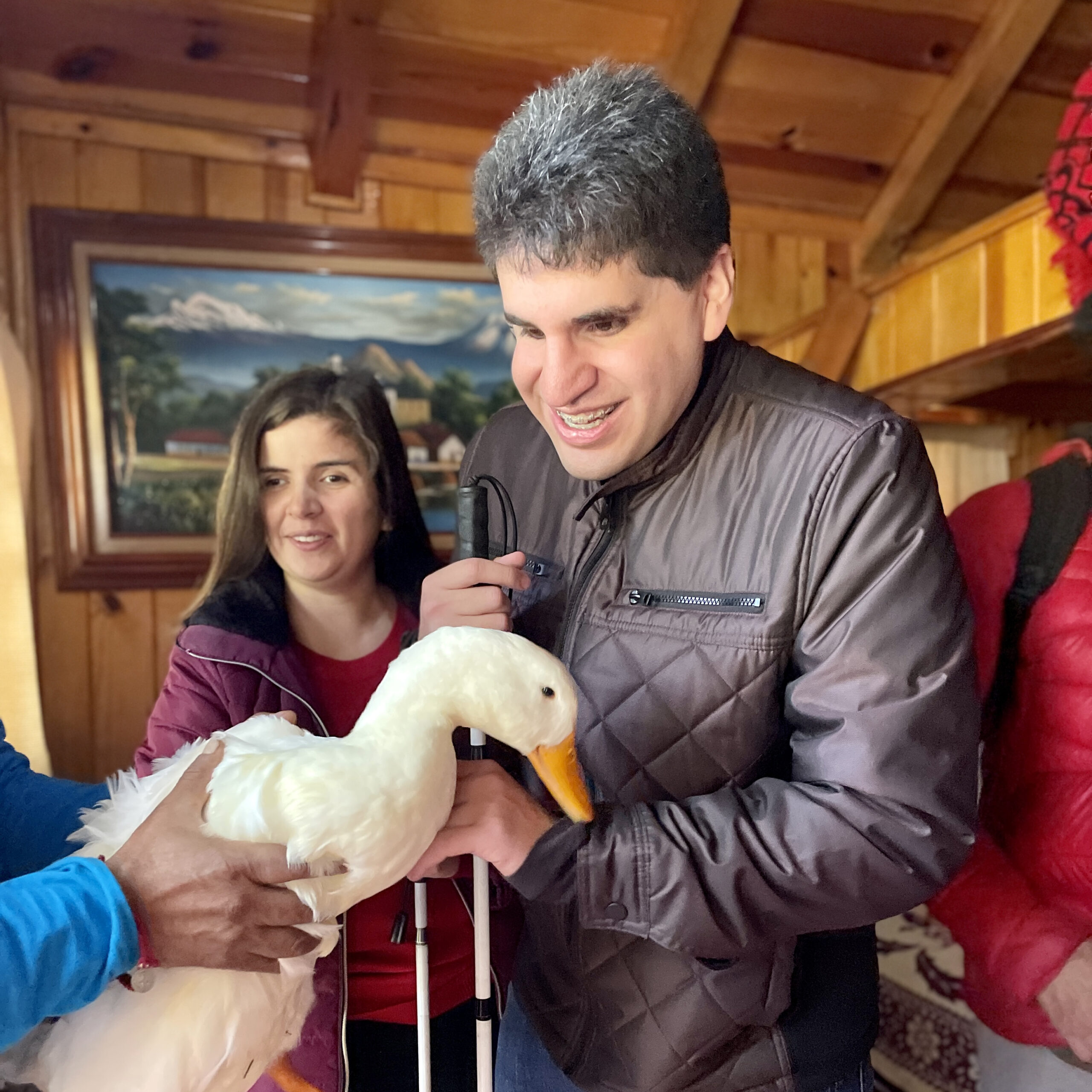 Fotografía cuadrada de una pareja con discapacidad visual dentro de una cabaña. A su lado izquierdo se aprecian las manos de una tercera persona cargando un pato. La pareja sonríe mientras acarician las plumas del pato.