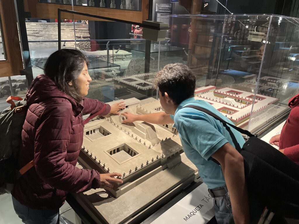 Fotografía de dos personas con discapacidad visual sintiendo con las manos una maqueta táctil de la edificación del Calmecac, una institución educativa prehispánica. La maqueta está sobre una mesa, dentro de una sala de exhibiciones.