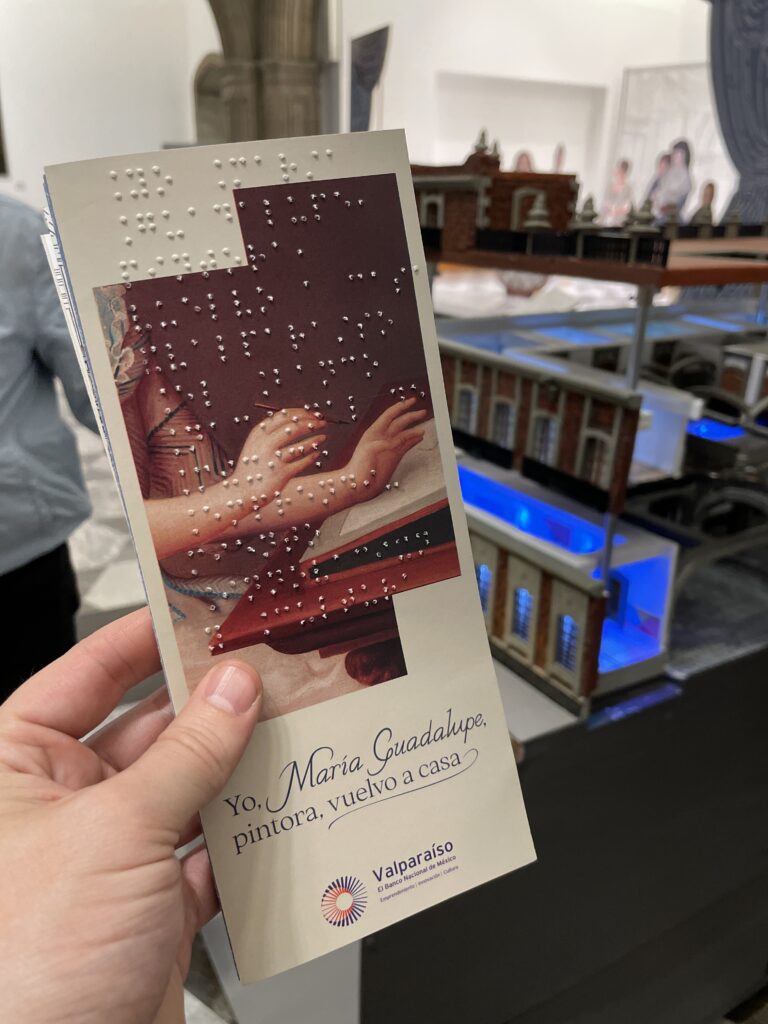 Fotografía vertical de un folleto con información impresa tanto en tinta como en Braille. El folleto lleva el título de la exposición "Yo, María Guadalupe, Pintora, vuelvo a casa". Al fondo se alcanza a ver una maqueta táctil del Foro Valparaíso, donde se encuentra la exposición.