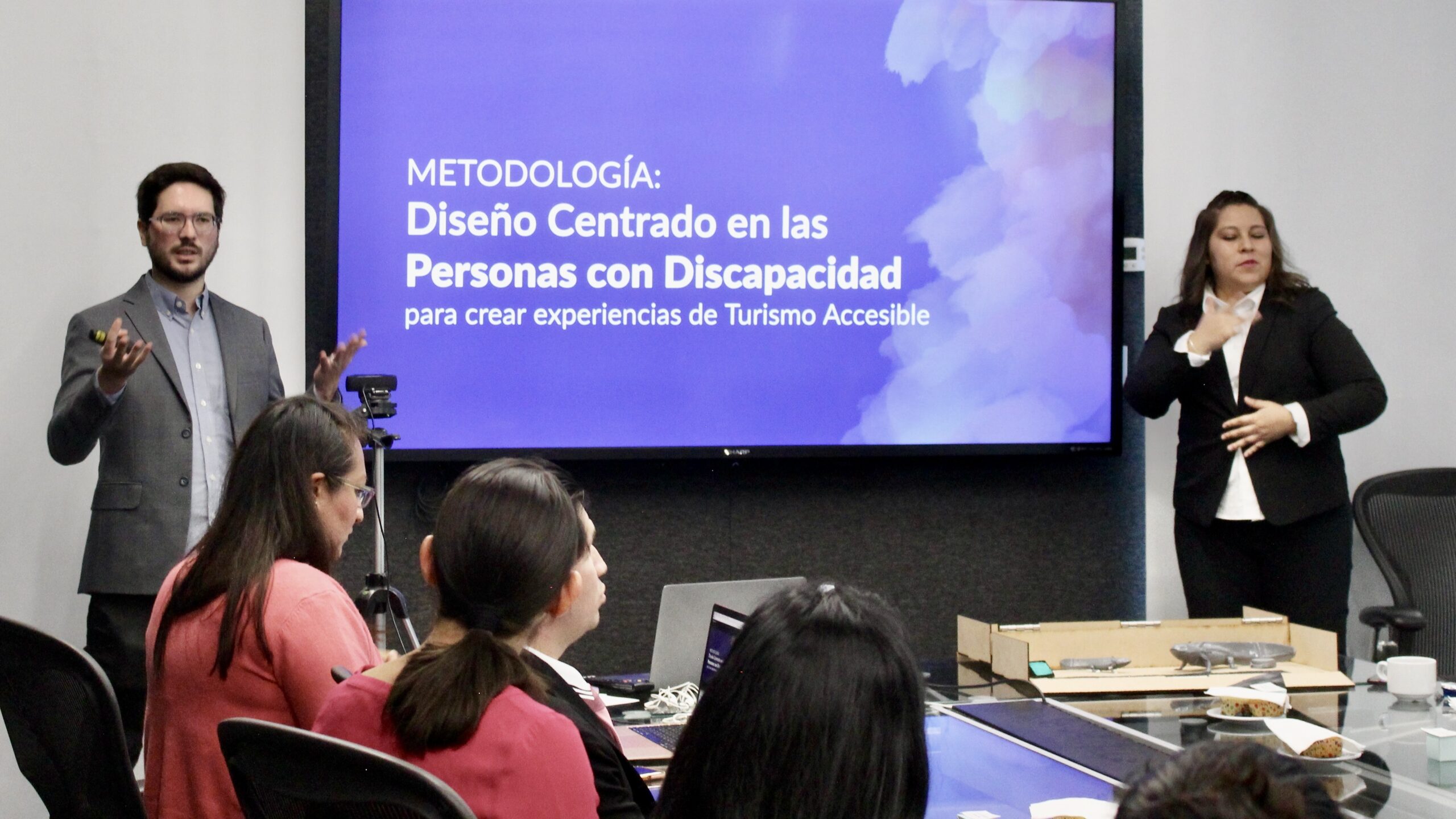 Fotografía de una persona dando una conferencia sobre "diseño centrado en las personas con discapacidad". A su derecha se encuentra una intérprete de lengua de señas mexicana. Se aprecian otras personas de espaldas, sentadas en el público.