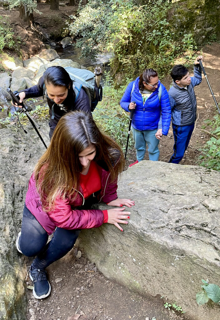 Fotografía de cuatro personas en una caminata en el bosque de los Dinamos. Al frente aparece una mujer con discapacidad visual trepando una roca. Detrás de ella se ubica una guía de turistas dándole indicaciones sobre cómo subir. Al fondo aparece una mujer sosteniendo el brazo de un hombre con discapacidad visual, que con un bastón está identificando las imperfecciones del camino.