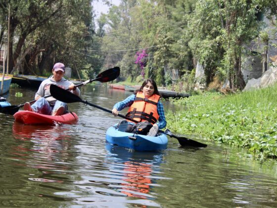 Fotografía de una turista con discapacidad visual en un kayak individual, remando por los canales de Xochimilco. Un guía le da indicaciones para saber hacia qué lado debe dirigirse.
