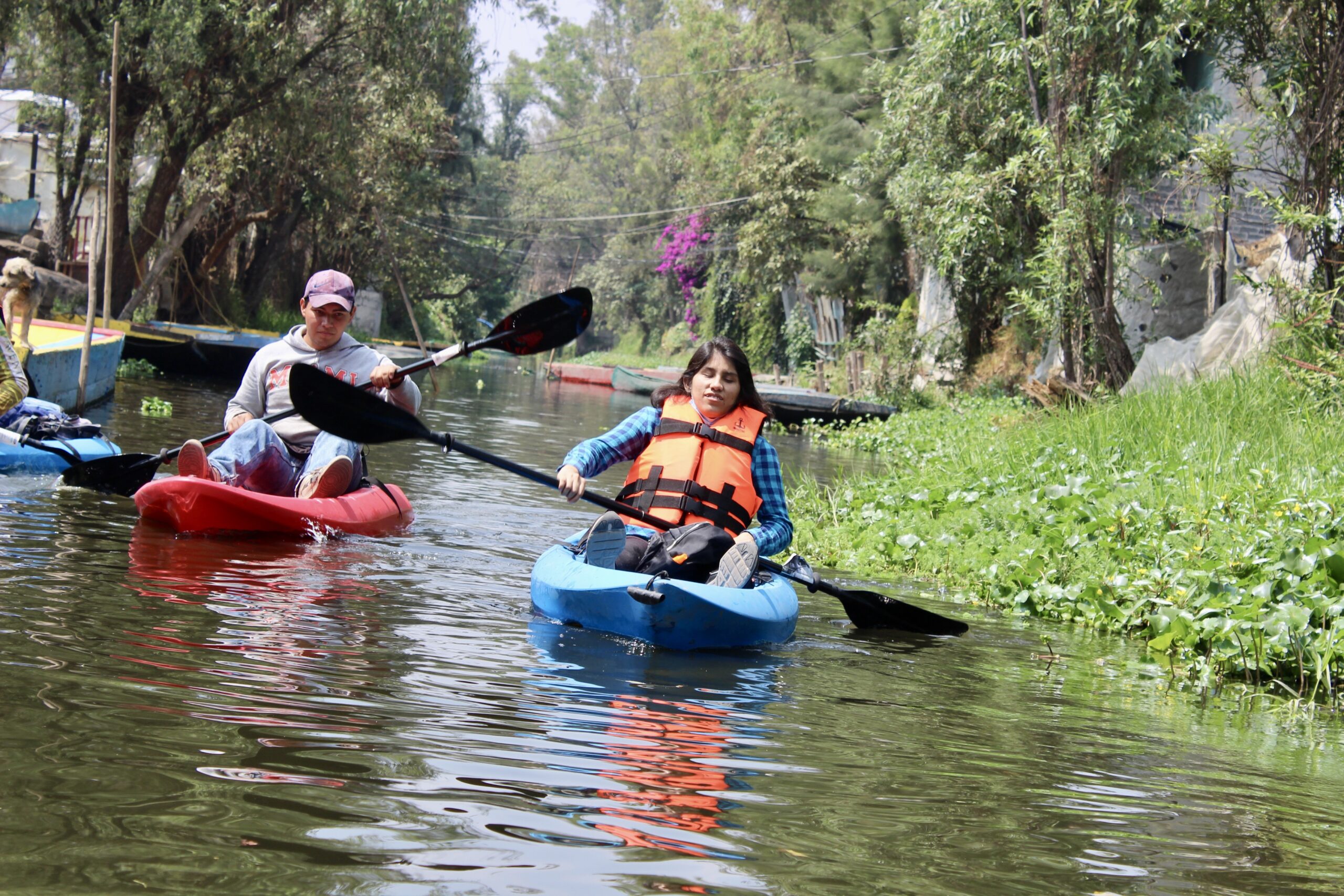 Fotografía de una turista con discapacidad visual en un kayak individual, remando por los canales de Xochimilco. Un guía le da indicaciones para saber hacia qué lado debe dirigirse.