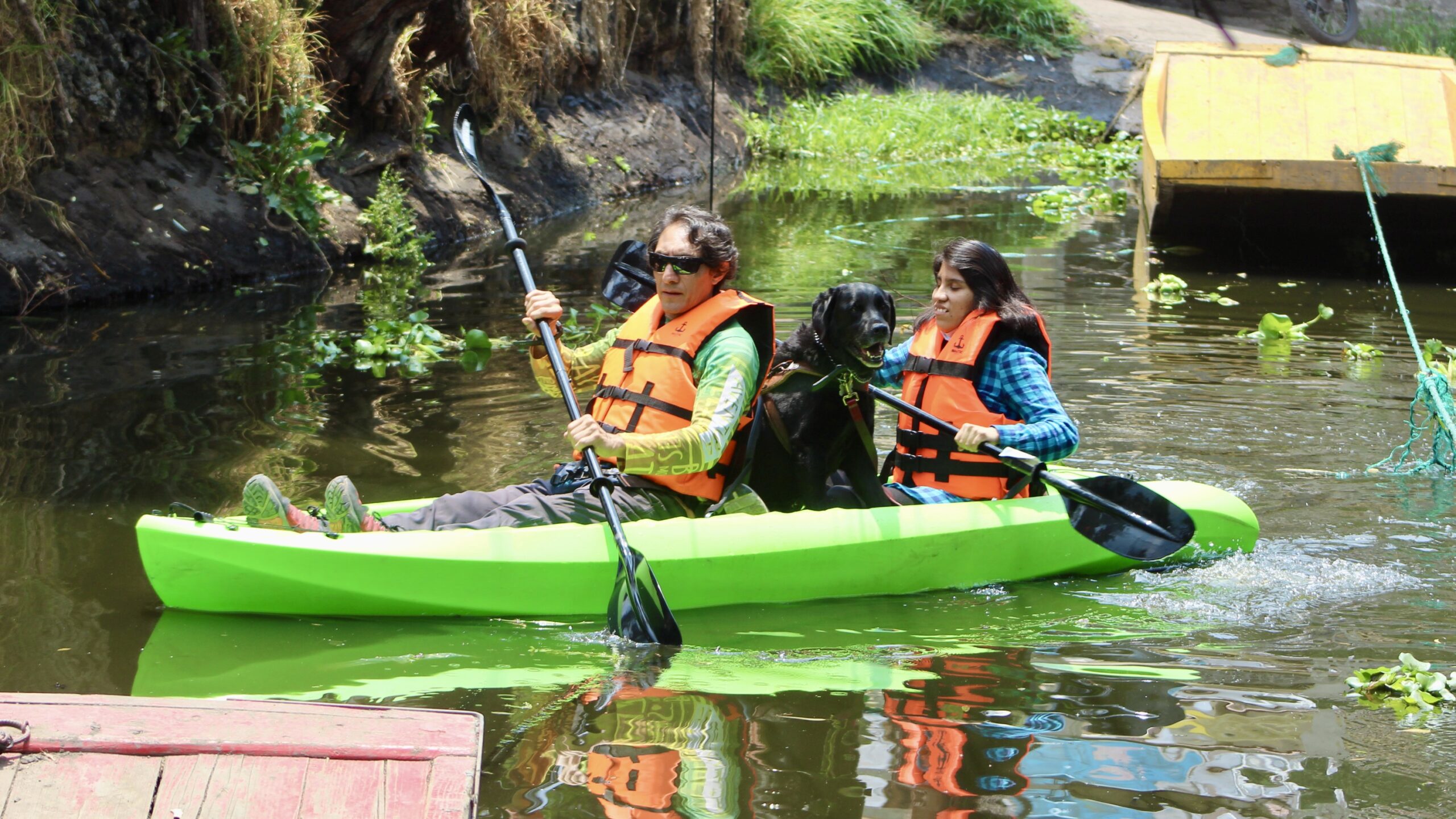 Fotografía de dos personas con remando en un kayak en los canales de Xochimilco. Una de ellas es persona con discapacidad visual, quien va a acompañada de un perro guía. Todos traen chaleco salvavidas.