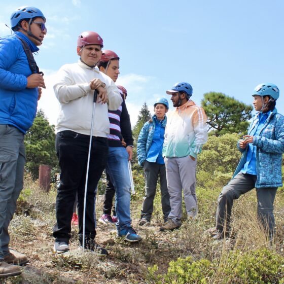 Fotografía horizontal de dos turistas con discapacidad visual con cascos de seguridad en la cima de una montaña, en el parque Natural el Rey. Están acompañados por guías, quienes les dan un recorrido de turismo accesible.