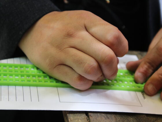 Acercamiento de las manos de una persona tratando de escribir su nombre en braille sobre un papel. Utiliza una regleta y un punzón.