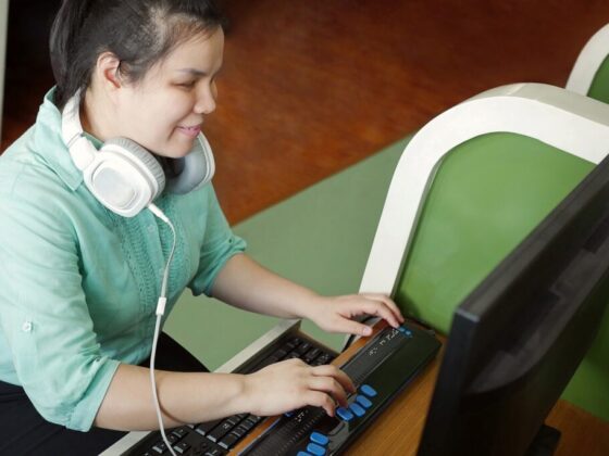 Fotografía de una mujer con discapacidad visual trabajando con una computadora. Usa audífonos para escuchar su lector de pantalla y escribe por medio de una línea braille.