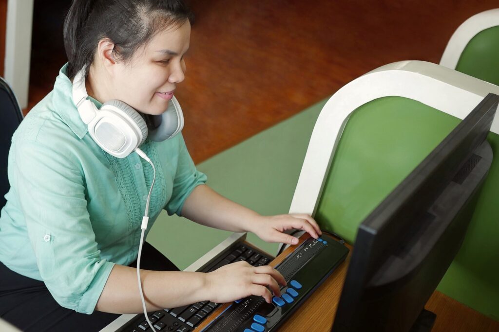 Fotografía de una mujer con discapacidad visual trabajando con una computadora. Usa audífonos para escuchar su lector de pantalla y escribe por medio de una línea braille.