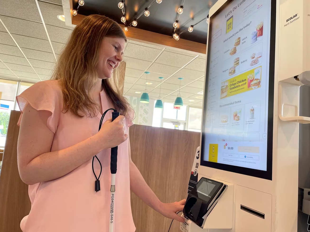 Fotografía de una mujer con bastón blanco ordenando comida en un módulo digital de McDonalds. Su mano está sobre un teclado a la altura de su abdomen, y escucha la información que está de forma visible en una pantalla vertical en la parte superior del módulo.