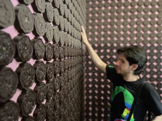 Fotografía de un hombre con discapacidad visual dentro de un cuarto tapizado por tablillas de chocolate. Las tablillas son circulares y están colocadas de piso a techo en los muros. El hombre extiende su brazo hacia arriba para tocarlas.