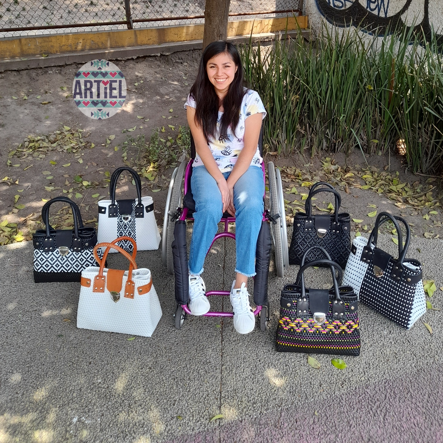 Foto de una mujer con discapacidad motriz que posa junto a bolsos con diferentes diseños hechos por ella, de la marca Artiel.mx