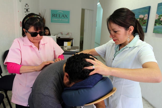 Fotografía de dos mujeres dando fisioterapia a un hombre recostado boca abajo. Una de las mujeres tiene discapacidad visual.