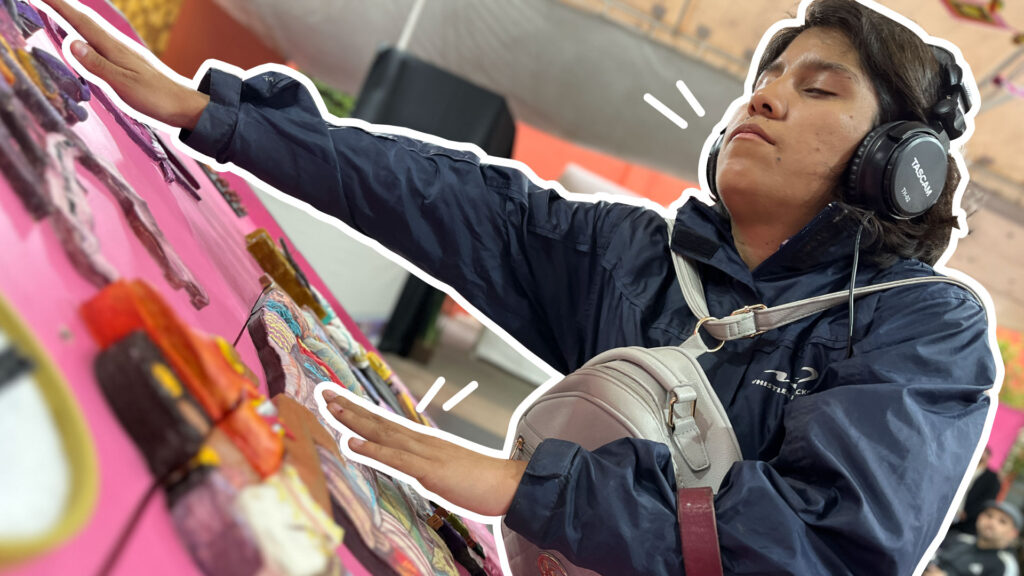 Foto de una mujer con discapacidad visual estirando los brazos para sentir las figuras en alto relieve con diferentes texturas, hechas para hacer accesible un mural. Están montados en una especie de mesa inclinada, y la mujer trae audífonos con los que escucha una descripción del mural.