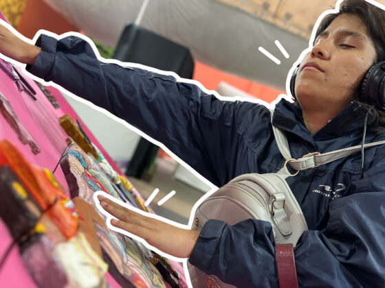 Foto de una mujer con discapacidad visual estirando los brazos para sentir las figuras en alto relieve con diferentes texturas, hechas para hacer accesible un mural. Están montados en una especie de mesa inclinada, y la mujer trae audífonos con los que escucha una descripción del mural.