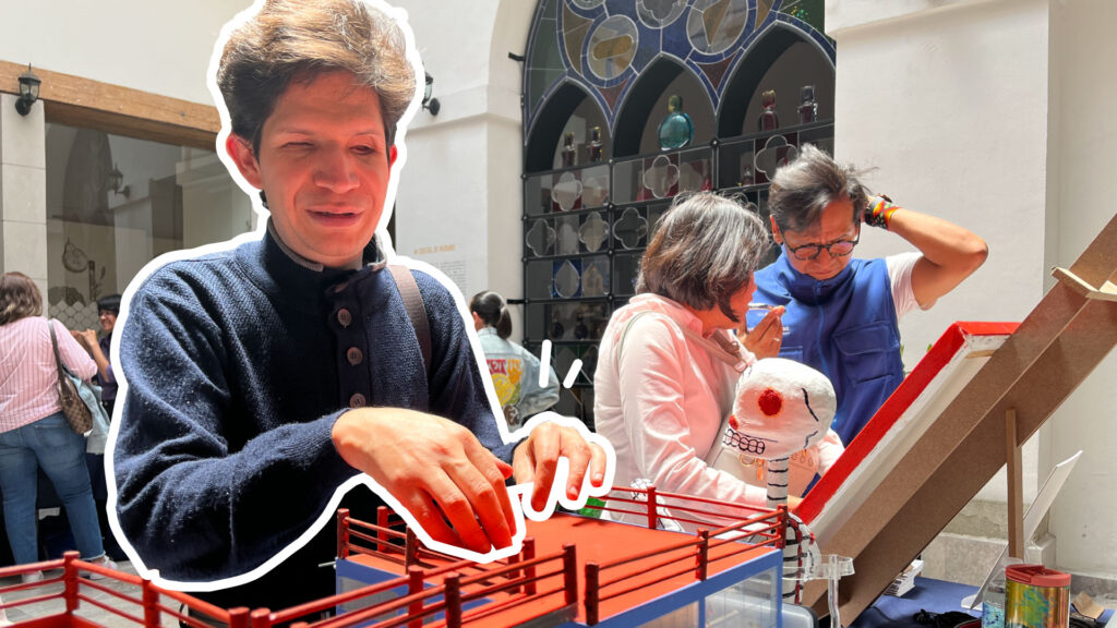 Foto de un hombre con discapacidad visual tocando una maqueta de la casa estudio Diego Rivera y Frida Kahlo. Atrás está una mujer con discapacidad visual tocando un cuadro con texturas en relieve. Un guía le intenta explicar de qué trata la obra, mientras se rasca la cabeza.