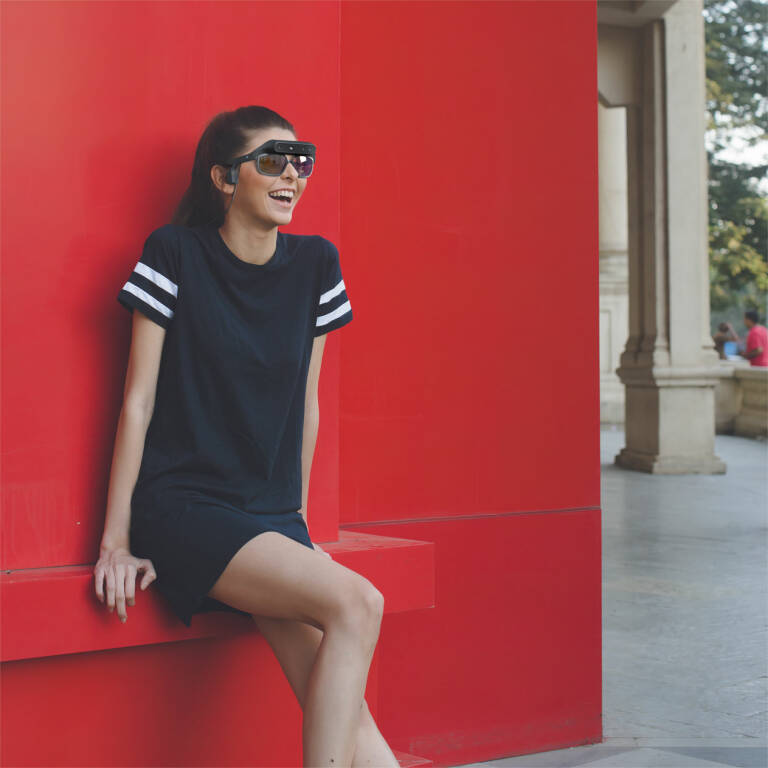 Foto de una mujer joven sonriendo, utilizando las gafas Niira. Está sentada en una banca delante de un muro rojo, en la calle.