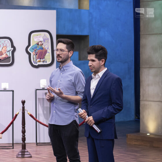 Foto de Alfonso y Cristóbal del IMETAC dando su pitch en el set de shark tank México. Cristóbal sostiene un bastón blanco en la mano. Alfonso levanta su mano mientras está hablando. Al fondo se ven unas piezas impresas en 3D.