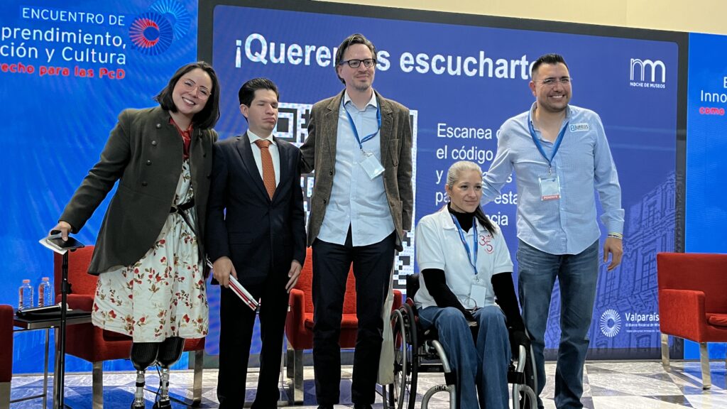 Foto horizontal de cinco ponentes en un escenario, posando para la foto. De izquierda a derecha: una mujer que usa prótesis en sus 2 piernas, un hombre con discapacidad visual de traje, un hombre con autismo con un saco, una mujer usuaria de silla de ruedas, y un hombre sin discapacidad.