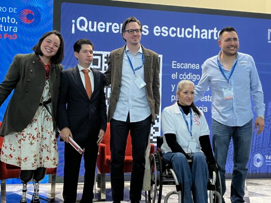 Foto horizontal de cinco ponentes en un escenario, posando para la foto. De izquierda a derecha: una mujer que usa prótesis en sus 2 piernas, un hombre con discapacidad visual de traje, un hombre con autismo con un saco, una mujer usuaria de silla de ruedas, y un hombre sin discapacidad.