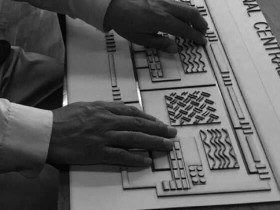 Foto vertical de un prototipo de mapa multimodal, formado por texturas como ondas, líneas en diferentes direcciones, letras y braille en alto relieve. Se aprecian las manos de una persona sintiendo las texturas.