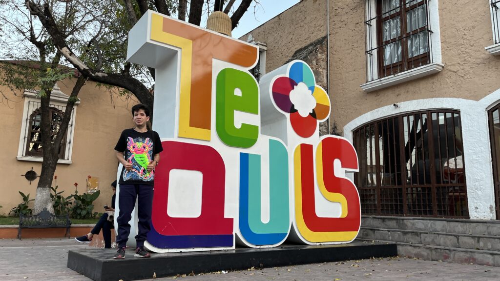 Foto de Cristóbal al lado de un letrero grande que dice Tequis, con letras más altas que él en diferentes colores.