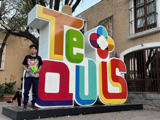 Foto de Cristóbal al lado de un letrero grande que dice Tequis, con letras más altas que él en diferentes colores.