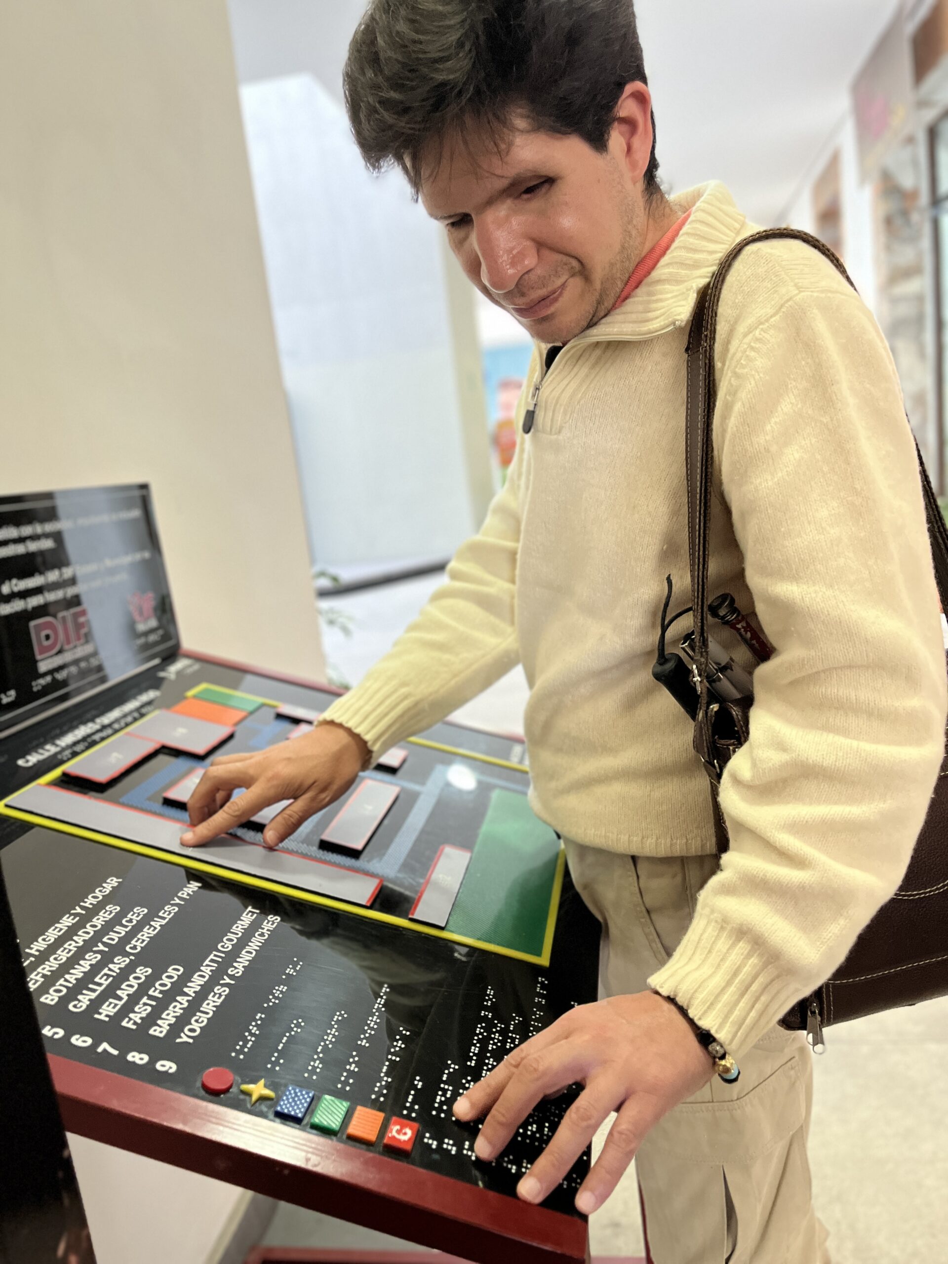 Foto de un hombre con discapacidad visual tocando el mapa háptico del Oxxo accesible. El mapa está en un pedestal metálico y cuenta con texturas y figuras en alto relieve para indicar cada espacio.