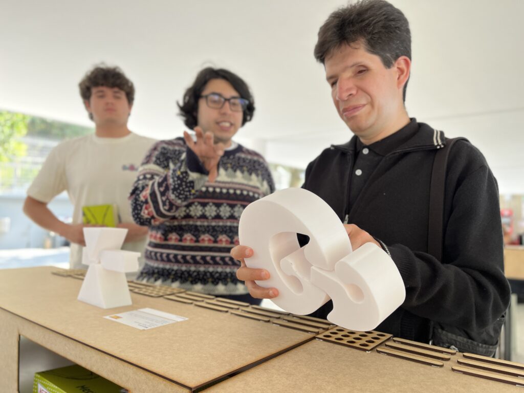 Foto de un hombre con discapacidad visual tocando una escultura a escala, impresa en 3D, que parece un ancla. Atrás se ven 2 estudiantes explicándole qué es.