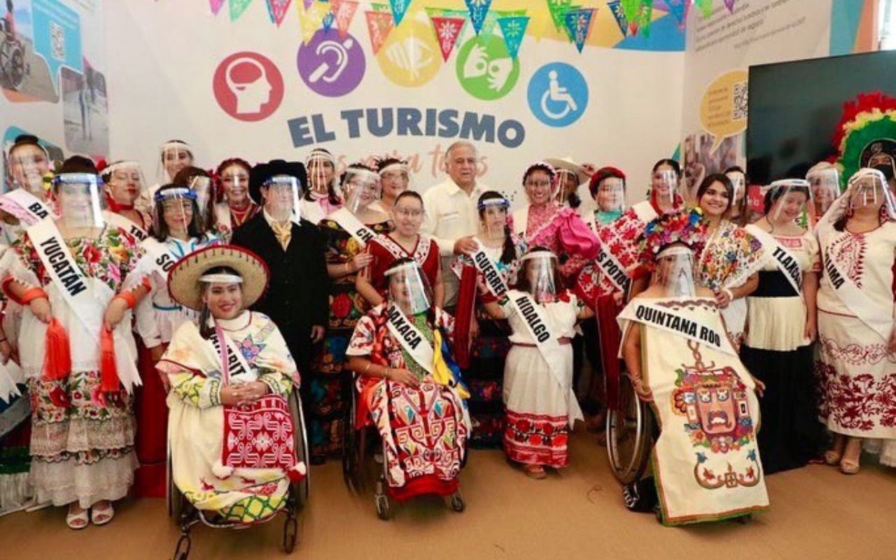 Foto de la inauguración del stand de turismo inclusivo de Tianguis Turístico 2021, en el que el ex secretario de turismo aparece posando para la foto con diversas mujeres con discapacidad que portan trajes típicos mexicanos.