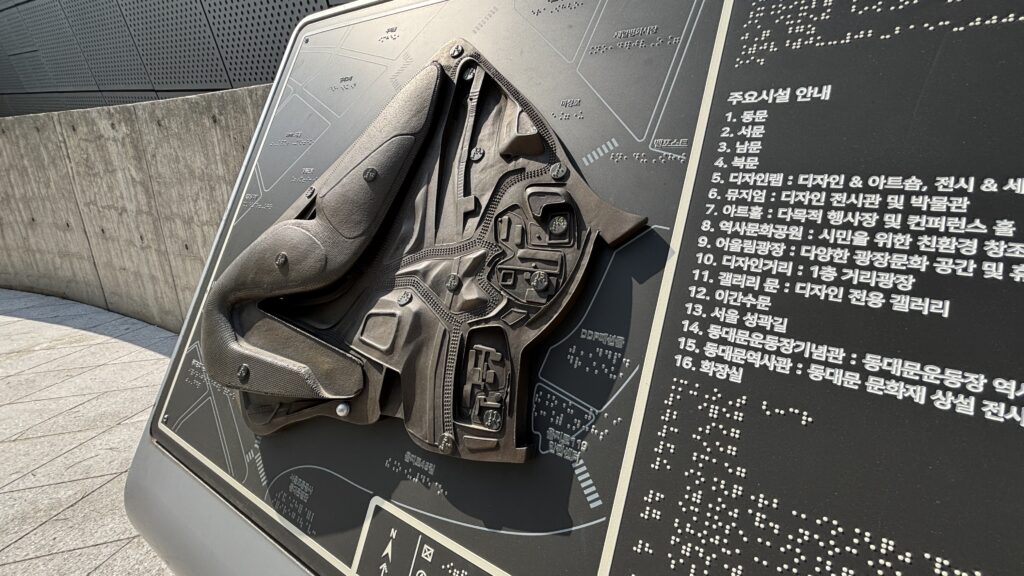 Foto horizontal de una maqueta de bronce de un edificio moderno, que parece una nave espacial. Está montada en un atril en posición vertical, y a su lado izquierdo aparece información en braille e impresa en coreano.