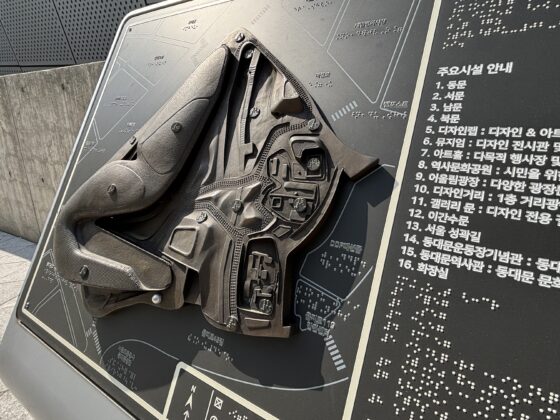 Foto horizontal de una maqueta de bronce de un edificio moderno, que parece una nave espacial. Está montada en un atril en posición vertical, y a su lado izquierdo aparece información en braille e impresa en coreano.