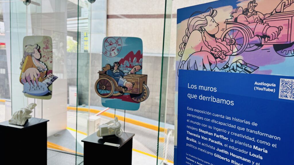 Foto horizontal de la exposición "los muros que derribamos" instalada en un corporativo. A la derecha, en primer plano se ve una mampara con el título de la exposición, un código QR para acceder a la audioguía y un texto. A la izquierda, más al fondo, se ven 2 módulos rectangulares sobre los que se exhiben dibujos de personajes y sus respectivas réplicas táctiles impresas en 3D.