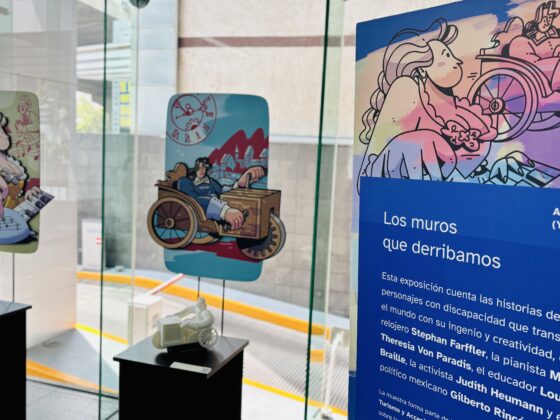 Foto horizontal de la exposición "los muros que derribamos" instalada en un corporativo. A la derecha, en primer plano se ve una mampara con el título de la exposición, un código QR para acceder a la audioguía y un texto. A la izquierda, más al fondo, se ven 2 módulos rectangulares sobre los que se exhiben dibujos de personajes y sus respectivas réplicas táctiles impresas en 3D.