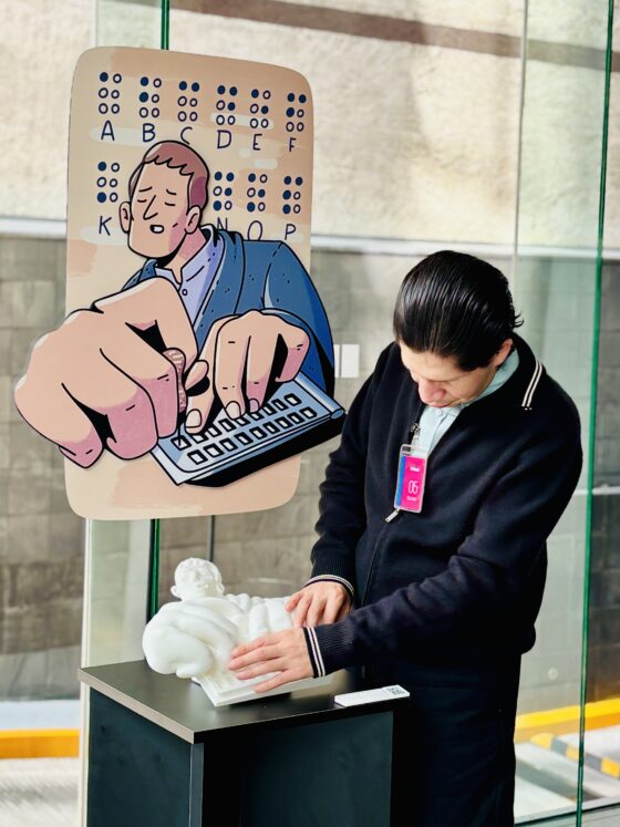 Foto vertical en perspectiva de un hombre con discapacidad visual tocando la réplica táctil del educador Louis Braille. La réplica está montada sobre un pedestal negro con una cédula en braille y un código QR, y al fondo se ve una ilustración de gran formato del mismo personaje.