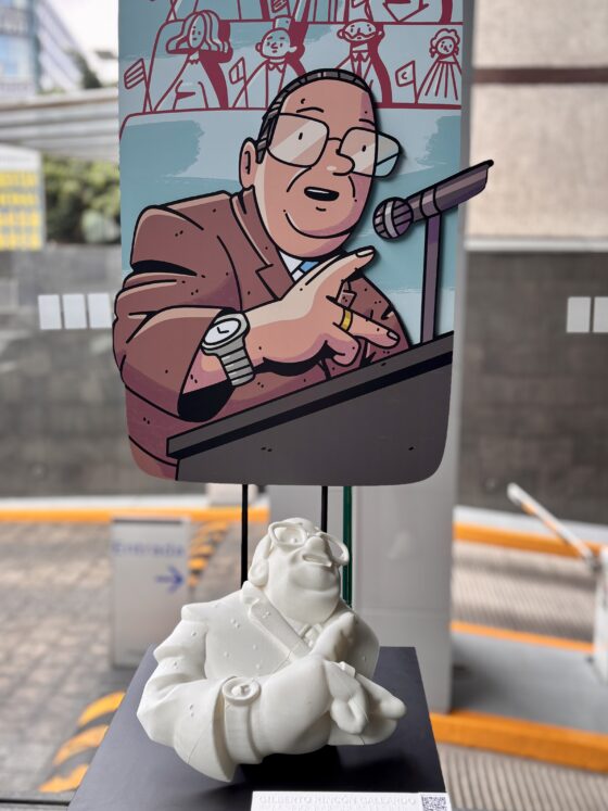 Foto vertical de un pedestal sobre el que se presenta al político mexicano Gilberto Rincón Gallardo, a través de una ilustración, una réplica táctil de la misma, y una cédula con información en braille y un código QR.